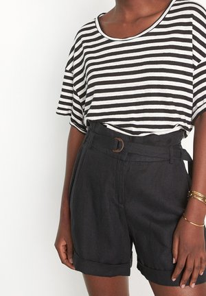 Person trägt schwarze hoch taillierte Shorts mit Gürtel und ein locker sitzendes schwarz-weiß gestreiftes kurzärmeliges Oberteil sowie goldene Armbänder am rechten Handgelenk.