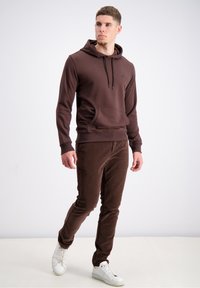 Brauner Hoodie mit einer Fronttasche und Kapuze mit Drawstring, kombiniert mit braunen Cordhosen und weißen Turnschuhen. Einfaches, lässiges Design.