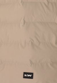 Beige vadderad jacka med quiltad textur, med horisontella linjer och en liten svart logotypapplikation märkt "SLOPE" på botten.