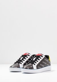 Des baskets noir et blanc arborent le logo "GUESS", avec des lacets noir et un accent jaune au niveau du talon. Texture lisse avec un design à tige basse.