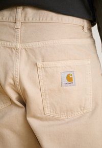 Pantalons Carhartt beiges avec une poche arrière dotée d'un patch logo cousu. Le tissu semble robuste avec des détails de couture visibles.