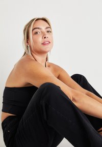 Jovem mulher com cabelo loiro, sentada de pernas cruzadas, vestindo um top preto, calças pretas e brincos pendentes, contra um fundo branco liso.