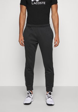 Man draagt een zwart Lacoste logo T-shirt, zwarte taps toelopende joggers met elastische boorden en witte sportsneakers met zwarte zolen.