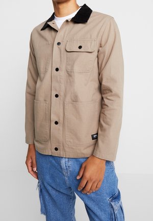 Homme portant une veste beige boutonnée avec un col noir et des poches avant, associée à un pantalon cargo en denim bleu.