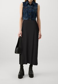 Denim ærmeløs jakke over en sort maxi-kjole. Sorte hælede støvler med kraftige såler og en sort håndtaske med en glat tekstur.