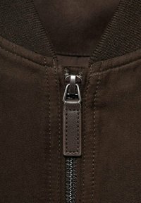 Braune Wildlederjacke mit einem Reißverschluss auf der Vorderseite, ausgestattet mit einem robusten Metallreißverschluss und einem gerippten Kragen. Der Stoff wirkt weich mit einem matten Finish.
