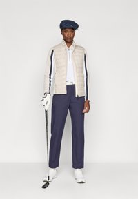Polstret kremfarget og marineblå golfvest over en hvit polo, marineblå bukser og hvite sneakers. Holder en golfkølle, har på seg en blå caps og hvite hansker.