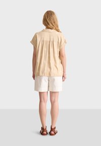 Blouse beige à manches courtes et col, avec un motif blanc subtil. Assortie à un short de couleur claire et des sandales brunes.
