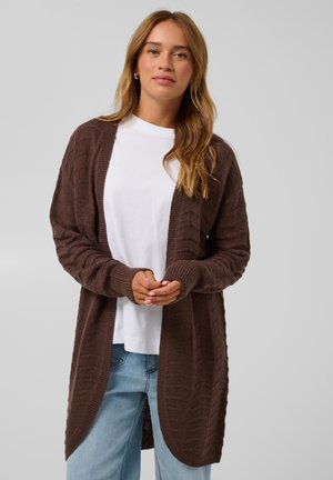 Frau mit hellbraunen Haaren, die ein weißes T-Shirt, blaue Jeans und einen langen braunen Strickcardigan trägt, steht vor einem schlichten Hintergrund.