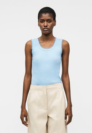 Μια γυναίκα στέκεται με το πρόσωπο προς τα εμπρός, φορώντας ανοιχτό μπλε αμάνικο τοπ με ξεφτισμένα άκρα και μπεζ παντελόνι με ψηλόμεση και φαρδιά μπάρα.