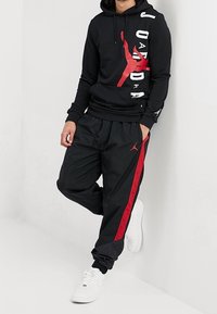Zwarte hoodie met rode Jordan-print, witte letters; zwarte trainingsbroek met rode streep, zwart-witte schoenen. De textuur lijkt glad.