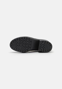 Semelle de chaussure noire avec des rainures profondes et géométriques ainsi qu’un talon texturé pour une bonne accroche, présentée sur un fond clair uni.