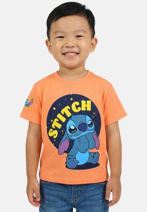 Jeune garçon souriant portant un t-shirt orange avec un grand personnage Stitch et du texte, associé à un jean bleu, sur un fond uni.