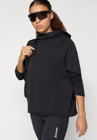 Μαύρος πουλόβερ με ψηλό λαιμό και μακριά μανίκια, διαθέτοντας ραφές λεπτομέρειες. Φορεμένο με ταιριαστά μαύρα κολάν και μεγάλα σκούρα γυαλιά ηλίου.