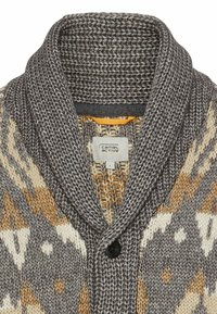Gestrickter grauer Cardigan mit einem Schalkragen, der ein geometrisches Muster in Beige und Weiß aufweist. Schwarzer Knopfverschluss und Etikett sichtbar im Inneren.
