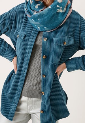 Personne portant une veste boutonnée en velours côtelé bleu, un pull gris côtelé, un pantalon blanc et une écharpe à motifs bleu sarcelle autour du cou.