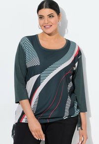 Blusa verde oscura con mangas tres cuartos, que presenta patrones geométricos en rojo, blanco y azul. Incluye detalles de cordón en los laterales.