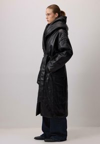 Sort læderlang frakke med bred hætte, bælte i taljen og to sidelommer. Glat tekstur med oversized design og afslappet pasform.