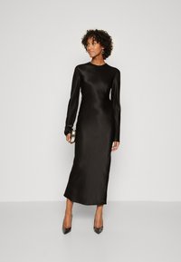 Svart satin langermet kjole med høy hals, midilengde og en glatt, shiny tekstur. Kombinert med spisse svarte hæler og en liten metallisk clutch.