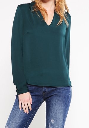 Bluzka z jedwabiu w kolorze morskim z dekoltem w kształcie litery V, długimi rękawami i zapinanymi na guziki mankietami, zestawiona z przetartymi niebieskimi dżinsami. Gładka faktura, prosty design.