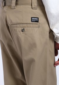 Beige broek met een fijne, gladde textuur. Voorzien van een achterzak met knoopsluiting en een zichtbaar merklabel dat op de zak is gestikt.