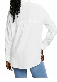 Chemise blanche à manches longues en tissu texturé, dotée d'un empiècement au dos et d'un ourlet légèrement arrondi, portée avec un jean bleu.