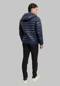 Giubbotto imbottito blu navy con cappuccio, caratterizzato da un design trapuntato orizzontale e una texture lucida. Abbinato a pantaloni neri e sneakers di colore chiaro.