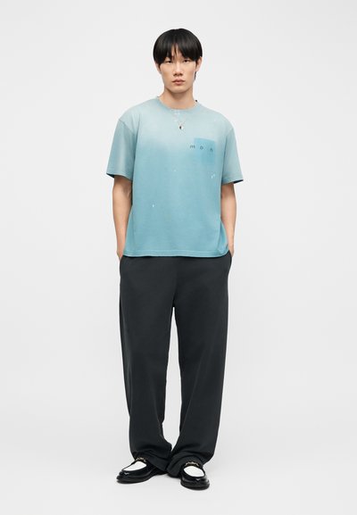 Νεαρός άντρας στέκεται με τα χέρια στις τσέπες, φορώντας ένα ανοιχτό μπλε φαρδύ μπλουζάκι, χαλαρά μαύρα παντελόνια και μαύρα-λευκά μοκασίνια.