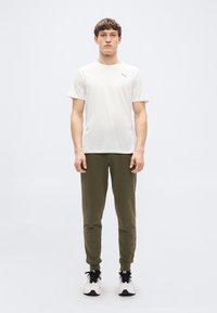 Λευκό αθλητικό t-shirt με κοντά μανίκια και μικρό λογότυπο, συνδυασμένο με χακί πράσινα παντελόνια τζόγκινγκ και ασπρόμαυρα αθλητικά παπούτσια.