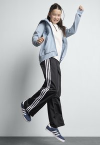 Blue zip-up hoodie over a white shirt, paired with black track pants featuring white stripes. Blue sneakers with white accents.