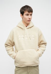 Νεαρός άνδρας που φοράει μια μπεζ φούτερ με γούνα και μπροστινή τσέπη, με το "JACK & JONES" κεντημένο στο στήθος, κοιτώντας στο πλάι.