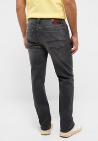 Szürke farmernadrág hátsó zsebekkel és bőr folttal a derékpánton. Testhezálló szabású, egyenes lábú, fehér sportcipővel viselve.