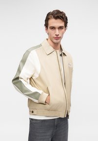 Beige und grüne Bomberjacke mit Reißverschluss, ausgestattet mit kontrastierenden weißen Ärmeln und gerippten Bündchen; Logopatch auf der Brust und zwei Seitentaschen.