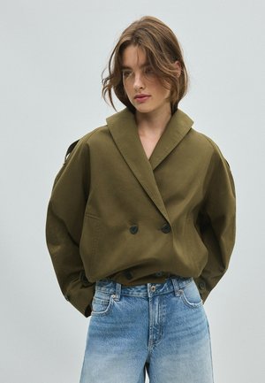 Jeune femme aux cheveux courts et bruns portant une veste oversize vert olive et un jean bleu clair sur un fond uni.