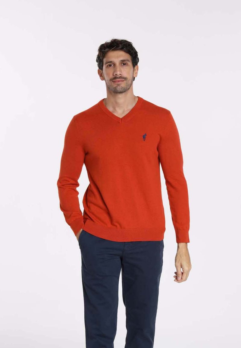 Pull-over orange en V fabriqué en tricot, avec des manches longues et des poignets côtelés. Détail du logo sur la poitrine gauche. Pantalons sombres portés en dessous.