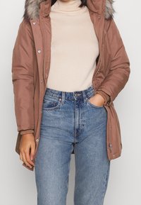 Brązowy zimowy parka z kapturem wyłożonym futrem, zamek z przodu, kieszenie po bokach i ściągacze na rękawach, w połączeniu z jasnoniebieskimi spodniami typu high-waisted i beżową bluzką.