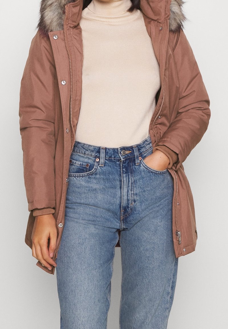 Brązowy zimowy parka z kapturem wyłożonym futrem, zamek z przodu, kieszenie po bokach i ściągacze na rękawach, w połączeniu z jasnoniebieskimi spodniami typu high-waisted i beżową bluzką.