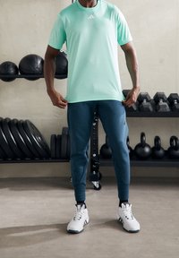 Person i mintgrön sporttröja och marinblå joggingsbyxor står i ett gym med vikter, kettlebells och medicinbollar i bakgrunden.