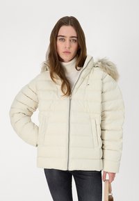 Chaqueta acolchada de un beige claro con cierre de cremallera, diseño acolchado y un ribete de piel sintética en la capucha. Presenta bolsillos laterales y un detalle de logo en el pecho.