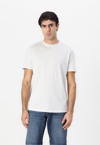Hvit bomulls t-skjorte med rund hals og korte ermer, prydet med en liten firkantet logo på venstre bryst, iført med denimshorts.