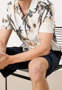 Camisa de manga corta, con cuello, en crema con patrones abstractos en verde, marrón y azul marino. Tela suave y cierre de botones en el frente. Pantalones cortos azul marino.