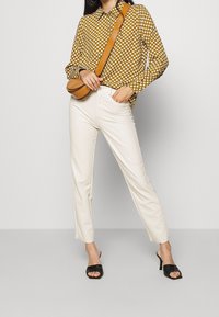 Blusa gialla a fantasia, pantaloni bianchi su misura e mule nere con tacco. Borsa a spalla in pelle marrone con forma curvata e tracolla.