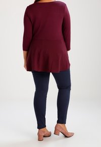 Burgunderfarbene Tunika mit dreiviertel Ärmeln, ausgestelltem Saum, kombiniert mit dunkelblauen Skinny Jeans und hellrosa Schuhen, von hinten betrachtet.
