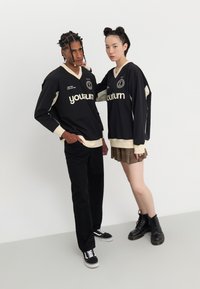 Jerséis negros de manga larga con acentos y logotipos en crema, combinados con pantalones negros y una falda de cuadros, botas de combate negras y zapatillas deportivas.