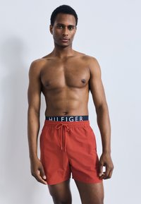 Κόκκινα μαγιό με κορδόνι, με σκούρο μπλε μέση και το κείμενο "HILFIGER". Το ύφασμα είναι λείο, κατάλληλο για δραστηριότητες στο νερό.