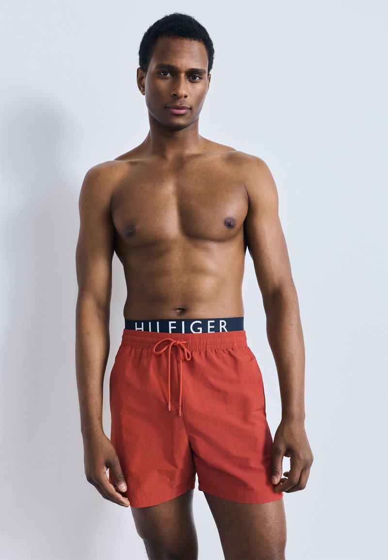 Κόκκινα μαγιό με κορδόνι, με σκούρο μπλε μέση και το κείμενο "HILFIGER". Το ύφασμα είναι λείο, κατάλληλο για δραστηριότητες στο νερό.