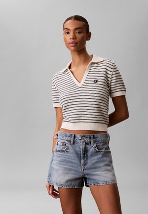 Frau trägt ein weiß-schwarzes gestreiftes Kurzarm-Stricktop und hellblaue Jeansshorts, steht vor einem grauen Hintergrund.