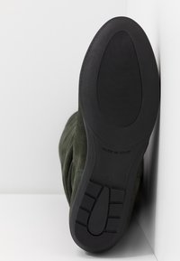 Vihreä mokkanahkasaapas, jossa on musta kumipohja, jossa on kuviollinen pohja ja sileä pinta, merkattu koolla 37 ja "VALMISTETTU ESPANJASSA."