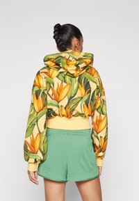 Geltona medvilninė cropped hoodie su gėlėta raštu, kuriame matosi oranžinės gėlės ir žalios lapijos. Derinama su žaliomis šortais.
