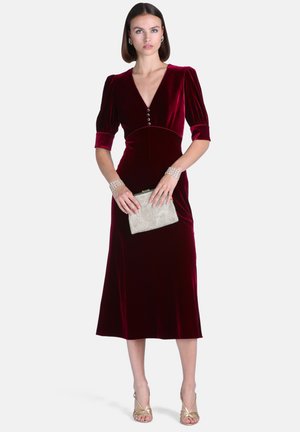 Femme portant une robe midi en velours bordeaux avec des manches bouffantes, tenant une pochette argentée, et portant des talons dorés à lanières et des bracelets ornés de bijoux.